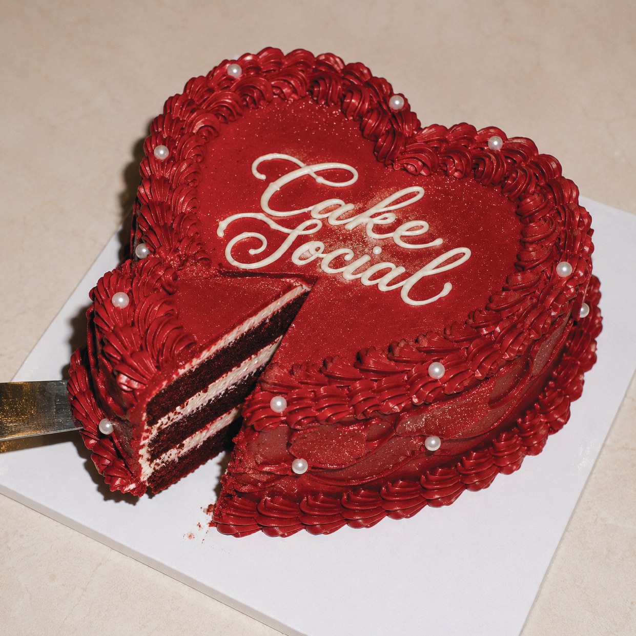 A deep red heart-shaped cake with Cake Social piped across it in white buttercream, pearls around the shell border and one slice cut away