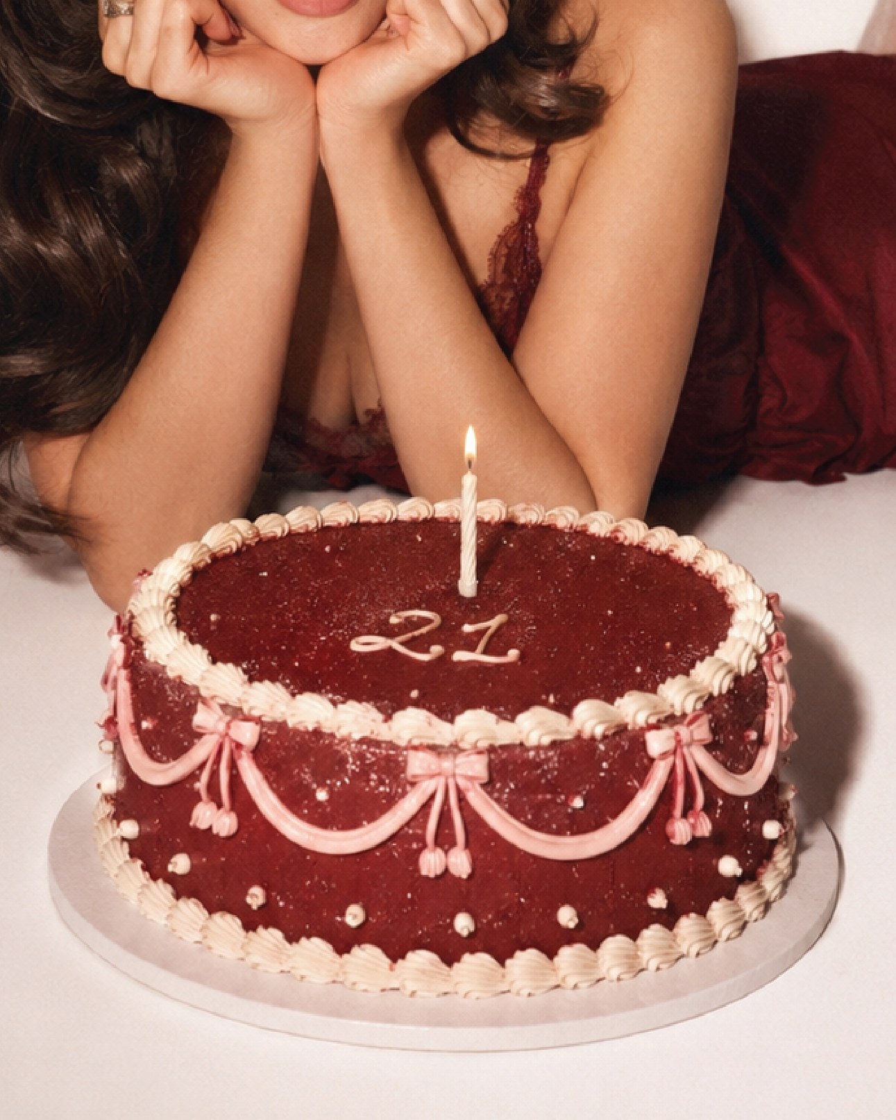 A woman in red lace resting her chin on her hands behind a red velvet cake with a single lit candle and a piped 21